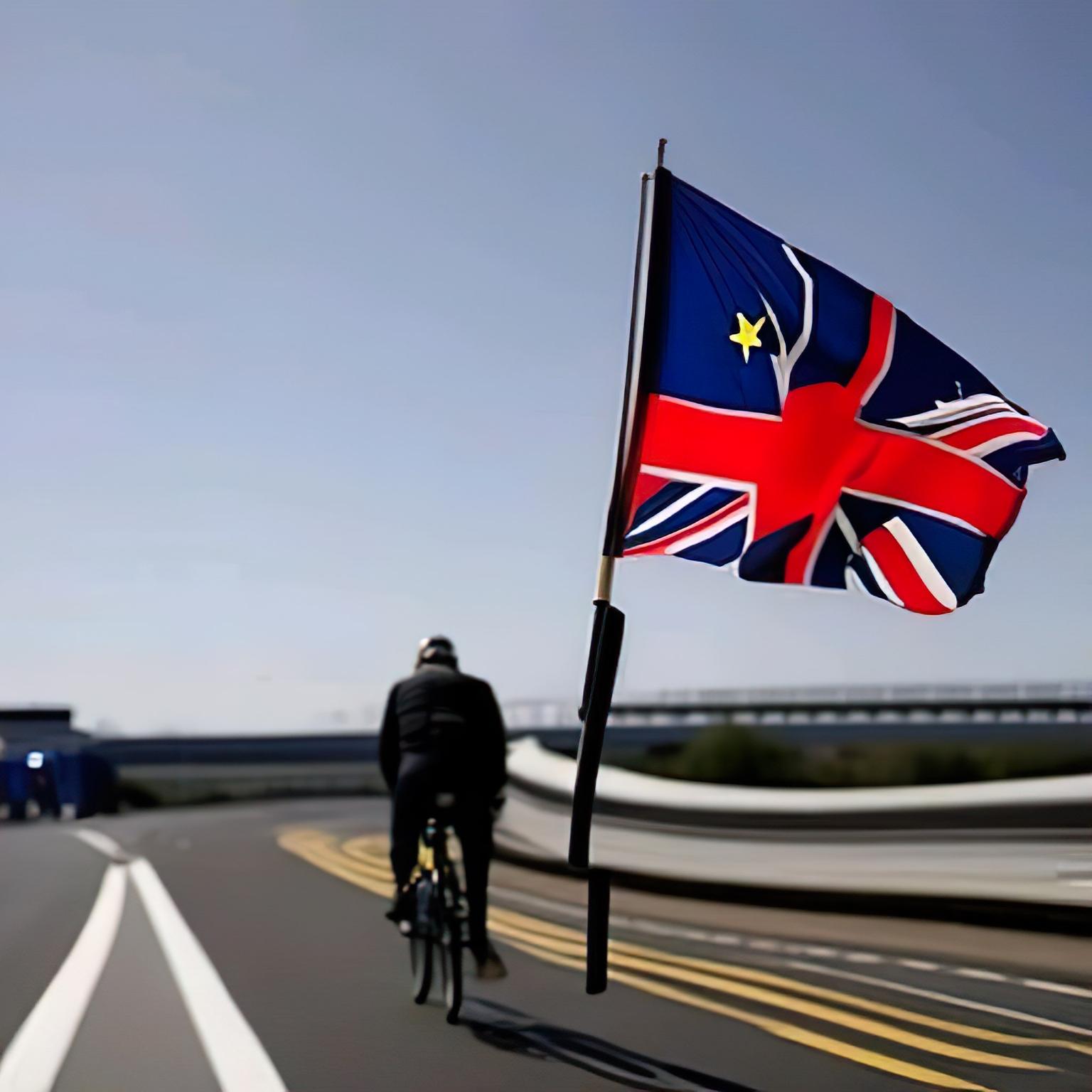 A man on a bicycle ride away on a European road with a floating corrupted union flag haunting him forever